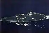 Large aircraft carrier with boxy island superstructure and numerous aircraft on its flight deck.