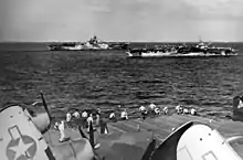 Black and white photo of two aircraft carriers sailing in close formation viewed from the flight deck of another aircraft carrier
