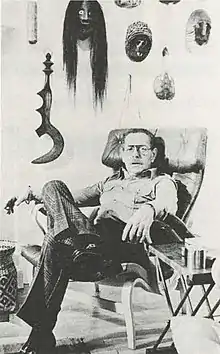 A white man with glasses seated with his legs crossed in front of a wall adorned with various masks and objects
