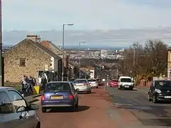 A busy main road with a distant view of an urban landscape. On the left are two-storey buildings; on the right are some mature trees.