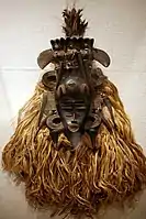 Poro mask; 19th-mid-20th century; wood, horns, raffia fiber, cotton cloth, feathers, metal; height: 30.mw-parser-output .frac{white-space:nowrap}.mw-parser-output .frac .num,.mw-parser-output .frac .den{font-size:80%;line-height:0;vertical-align:super}.mw-parser-output .frac .den{vertical-align:sub}.mw-parser-output .sr-only{border:0;clip:rect(0,0,0,0);clip-path:polygon(0px 0px,0px 0px,0px 0px);height:1px;margin:-1px;overflow:hidden;padding:0;position:absolute;width:1px}1⁄4 in.; by Senufo people; Metropolitan Museum of Art. Designed to pay homage to female ancestors, this mask's serene dark oval face is offset by glinting brass, symmetrical extensions, and delicate patterns symbolizing wisdom and beauty