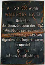 Damaged metal plaque reading "Am 3.9.1956 wurde WALDEMAR ESTEL Getreiter der Grenztruppen der NVA in Ausübung seine Dienstes an der Staatsgrenze von Agenten des Imperialismus ermordet. Sein Tod ist uns Verpflichtung."