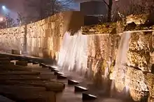 To the right of some stepping stones, a row of lamps illuminates a short artificial waterfall streaming down a honey-colored wall made of limestone bricks.