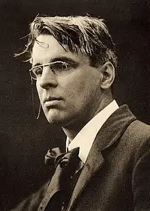 A posed black and white photograph of Yeats. He is wearing smart clothes and spectacles, while his hair looks a bit tousled