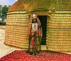 Woman in traditional dress standing on rug in front of yurt, circa 1905-1915