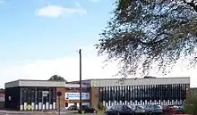 A building approximately 3 stories high. For the most part, the building is made up of horizontal columns of an opaque white panel up to around head height, with 3 windows arranged vertically above this. The columns are approximately 0.5&nbsp;m (1.6&nbsp;ft) wide and run from just below the roof to just above ground level and are separated by vertical wooden beams. Above the columns of windows a corrugated white border runs along the top of the building.