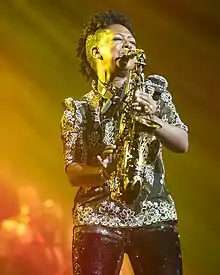A Black woman playing the saxophone with eyes closed. Lit from the side with yellow and lit overall in white, she is wearing a black shirt with floral patterns in white and green embroidery and glittery black trousers with Natural hair in a wide, short mohawk, shaved on either side. Behind her, some instruments are out of focus.