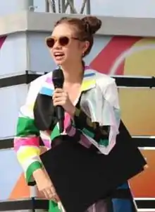 young Asian woman standing onstage, holding a microphone, wearing sunglasses and a patterned shirt