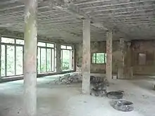 An empty room in an abandoned building with no finishes, cement structural systems visible, support columns in the middle, empty openings along a left brick wall through which a forest can be seen, and cement blocks and tires piled on the floor