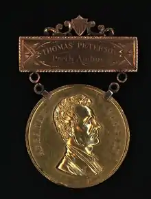 The engraving on the top bar reads "THOMAS PETERSON, Perth Amboy". The hanging medallion is attached to the top bar using two chains. The hanging medallion shows a profile bust of a clean-shaven Abraham Lincoln. The medallion awarded to Thomas Mundy Peterson by the citizens of Perth Amboy in 1884.