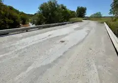 A short and unmarked concrete road bridge, with white gravel scattered atop, spans a brown creek with trees and grasslands on either side; graffito on the visible guardrail says, "GONE NOT FORGOTTEN ♥ RIP HEATHER 1996 ♥"