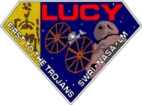 A diamond-shaped crest houses artworks of the Lucy fossil at left, the Lucy spacecraft at center, and an artist's impression of a Jupiter trojan. The word "Lucy" is written in a large, bold red font at the top right corner, while the words "First to the Trojans" and "SWRI&nbsp;· NASA&nbsp;· LM" are written in a smaller white font across the bottom edges of the diamond-shaped crest.