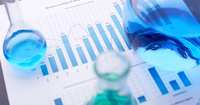 Photo showing a scientific report that contains two vertical bar graphs. Placed on top of the report are three volumetric flasks. From left to right the flasks contain a light blue liquid, a green liquid, a blue liquid. 