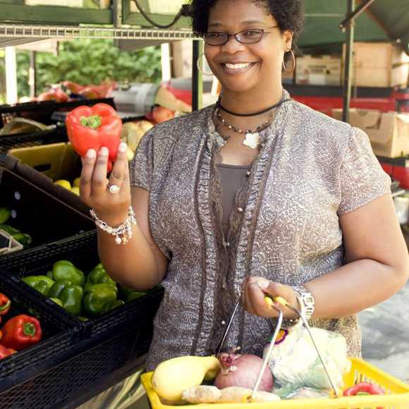 The photograph shows a women with her shopping basket filled with an array of healthy food choices, this woman had completed her selection process at a mobile produce market, and was shown holding up a bright red bell pepper. Some of the vegetables she’d placed into her basket included a lemon, a head of cauliflower, a purple and a white onion, ginger root, and a variety of bell peppers.