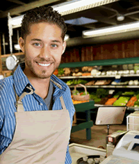 A young man wearing an apron stands near a cash register in a market.