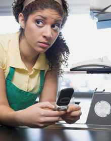 A young woman leans over a store counter where she works. She is holding a cell phone, staring off into the distance, looking frightened.